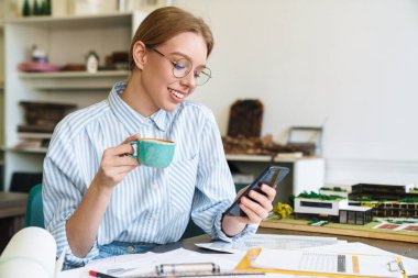 Mutlu fotoğrafçı kadın mimar kahve içerken ve cep telefonu kullanırken iş yerinde tasarım yaparken.