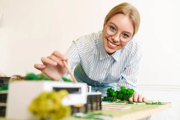 Photo of joyful young woman architect in eyeglasses designing draft with house model at workplace