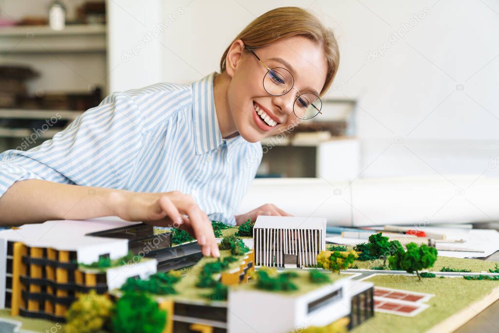 Photo of joyful young woman architect in eyeglasses designing draft with house model and sitting at workplace