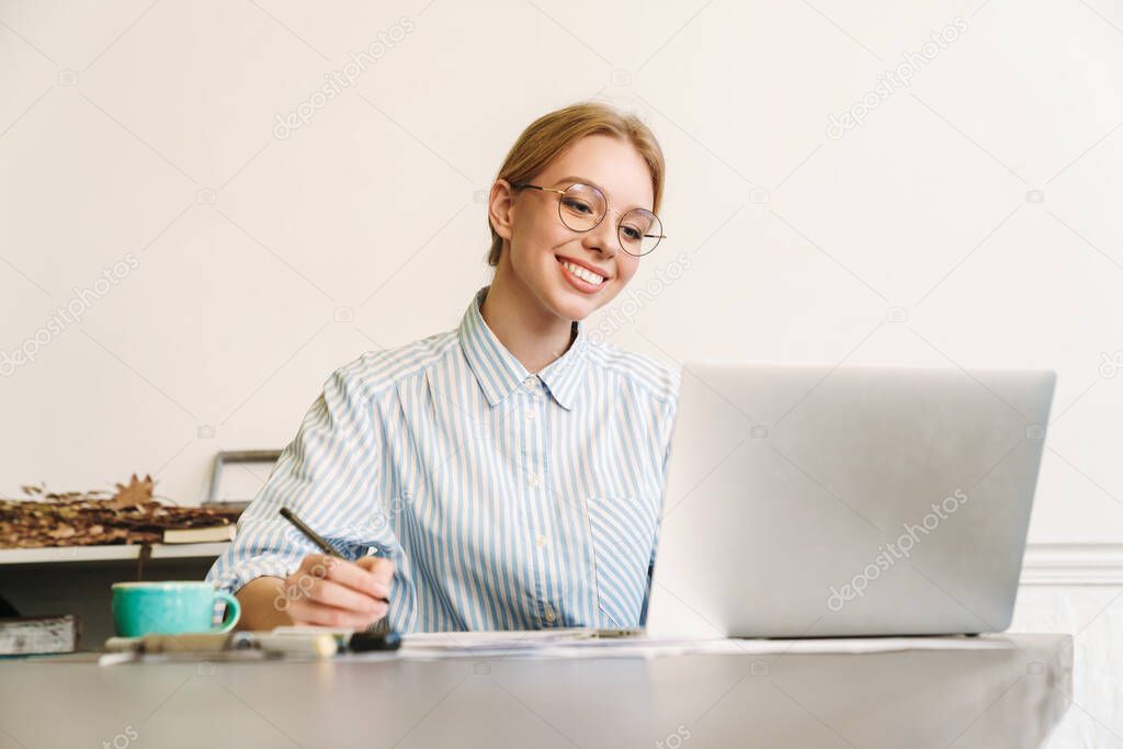 Photo of smiling blonde woman architect in eyeglasses working with laptop while designing draft at workplace