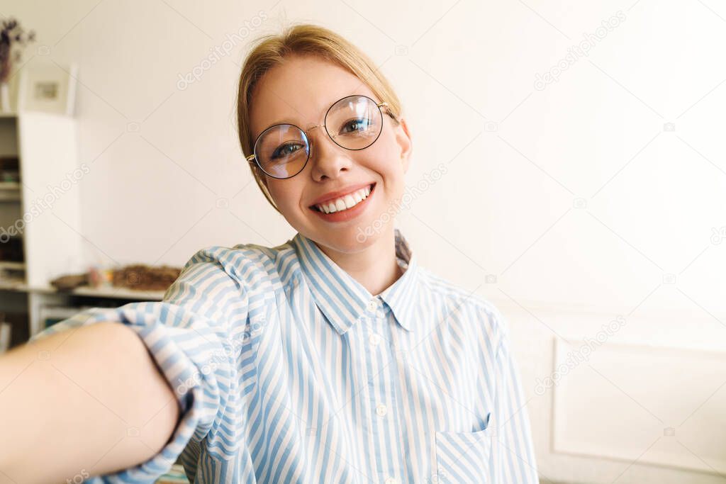 Photo of happy blonde woman wearing eyeglasses smiling while taking selfie photo in office