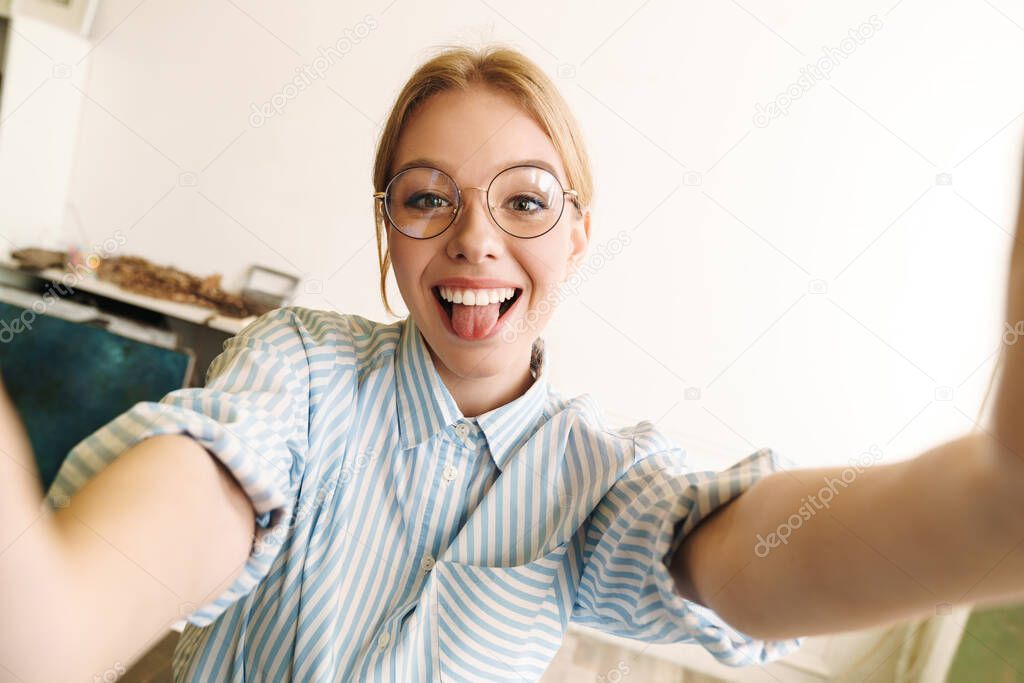 Photo of joyful blonde woman in eyeglasses sticking out her tongue while taking selfie photo at office