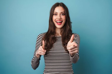 Mavi arka planda izole edilmiş, gülümseyen ve parmaklarını kameraya gösteren heyecanlı beyaz kadın görüntüsü