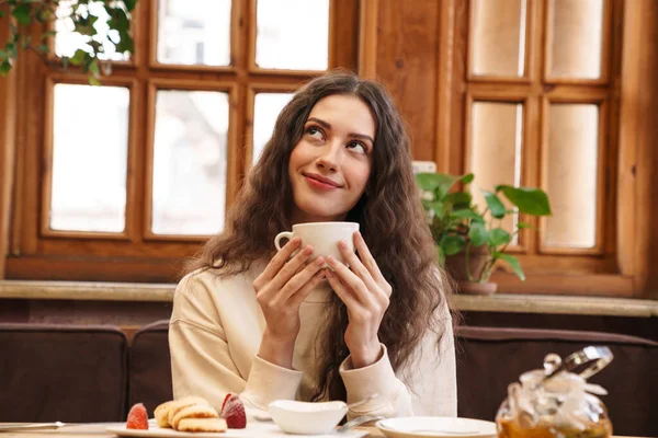 照片中快乐美丽的年轻女子坐在舒适的室内咖啡馆里 微笑着喝茶 — 图库照片