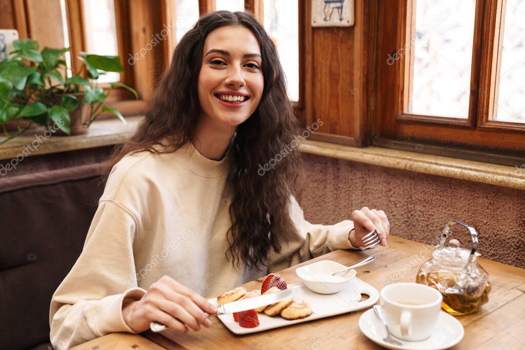 Image of happy beautiful young woman smiling and eating pancakes while sitting in cozy cafe indoors