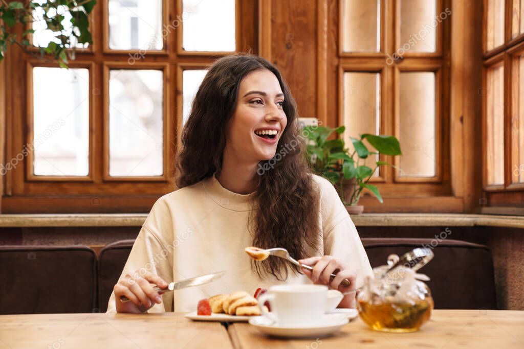 Image of happy beautiful young woman smiling and eating pancakes while sitting in cozy cafe indoors