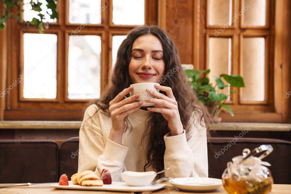 Image of happy beautiful young woman smiling and drinking tea while sitting in cozy cafe indoors