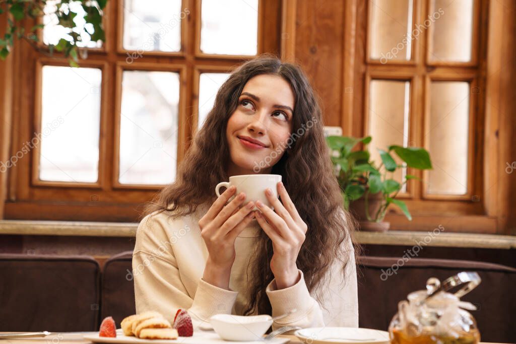 Image of happy beautiful young woman smiling and drinking tea while sitting in cozy cafe indoors