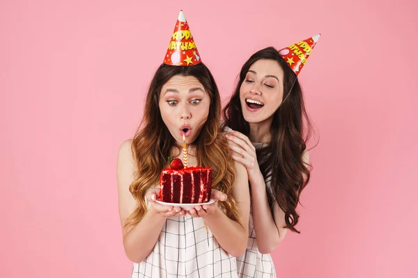 Image of excited beautiful women in party cones blowing out candle and smiling isolated over pink background