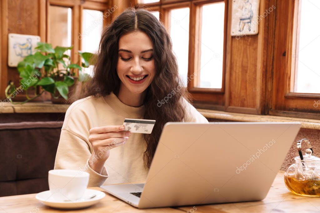 Image of smiling beautiful woman using laptop and holding credit card while drinking tea in cafe indoors