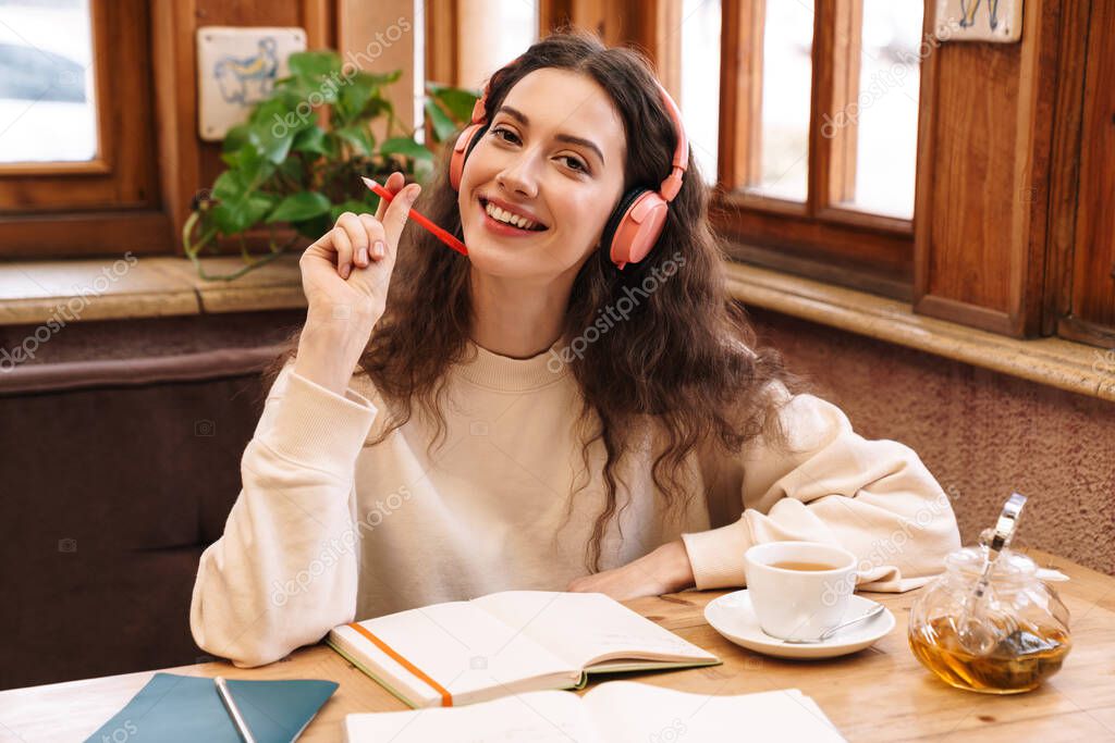 Image of joyful student woman using wireless headphones while doing homework in cozy cafe indoors