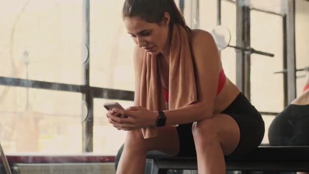 Joven Mujer Fitness Optimista Concentrado Gimnasio Utilizando Teléfono Móvil Mirando — Vídeos de Stock