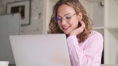Gözlüklü çekici sarışın bir kadın laptopunu kullanarak çalışıyor ve masada oturuyor.