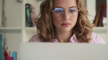 Düşünceli, hoşnutsuz ve ciddi, gözlüklü genç bir sarışın laptopunu kullanarak çalışıyor.