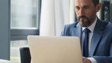 Ciddi bir yetişkin adam ofiste dizüstü bilgisayarıyla çalışıyor.