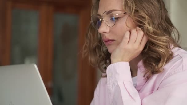 ガラスを身に着けている思慮深い深刻な若いブロンドの女性の側面図は 屋内のテーブルに座っている彼女のラップトップコンピュータを使用して作業しています — ストック動画