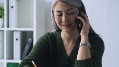 Olgun bir kadın ofiste çalışırken akıllı telefonla konuşurken notlar alıyor.