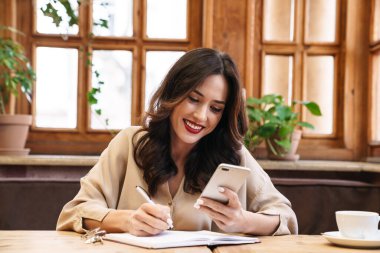 Gülümseyen yetişkin bir kadının günlük tutarken cep telefonunu kapalı bir kafede otururken kullanması.