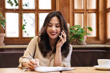 Gülümseyen yetişkin bir kadının günlük tutarken cep telefonuyla konuşurken kapalı bir kafede oturması.