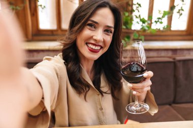 İçeride rahat bir kafede oturmuş şarap kadehiyle selfie çeken heyecanlı yetişkin bir kadın resmi.