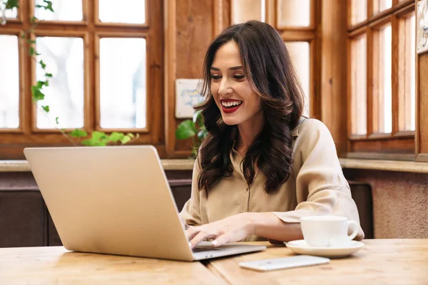 幸せなブルネットの大人の女性の笑顔と仕事ノートパソコンで座っている間居心地の良いカフェ室内 — ストック写真