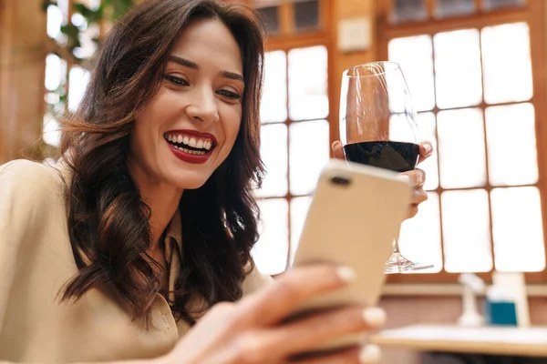 Kırmızı şarap içen ve kapalı bir kafede otururken cep telefonu kullanan neşeli yetişkin bir kadın.
