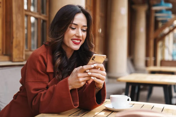 照片中 一个漂亮的成年女子坐在街上的咖啡店里 一边用手机一边喝咖啡 — 图库照片