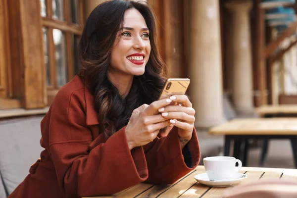 屋外のストリートカフェに座っている間 携帯電話や飲酒コーヒーを使用して美しい大人の女性を笑顔のイメージ — ストック写真
