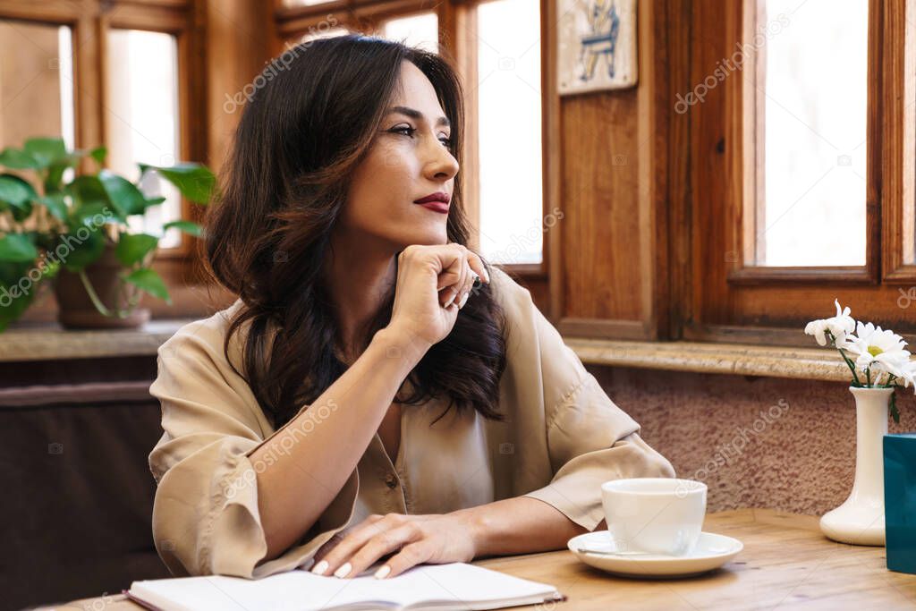 Image of thinking adult woman making notes in diary and drinking coffee while sitting in cozy cafe indoors