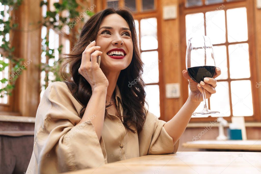 Image of cheerful adult woman drinking red wine and talking on mobile phone while sitting in cafe indoors
