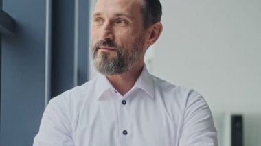 Çekici, olgun bir adam, ofiste çapraz ellerle pencerenin yanında dururken dışarıya bakıyor.