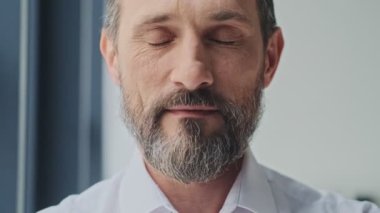 Sakin ve olgun bir adamın yakın çekim görüntüsü ofiste dikilirken gözlerini açar ve kameraya bakar.