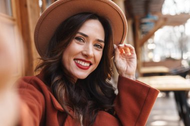 Şapkalı, neşeli, zarif, yetişkin bir kadının sokak kafesinde otururken gülümseyip selfie çektiği bir resim.