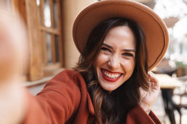 Şapkalı, neşeli, zarif, yetişkin bir kadının açık hava kafede otururken fotoğraf çekmesi.