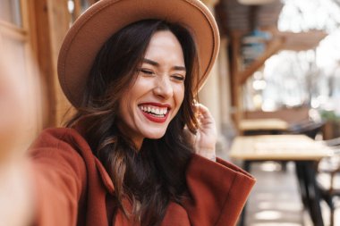 Şapkalı, neşeli, zarif, yetişkin bir kadının açık hava kafede otururken fotoğraf çekmesi.