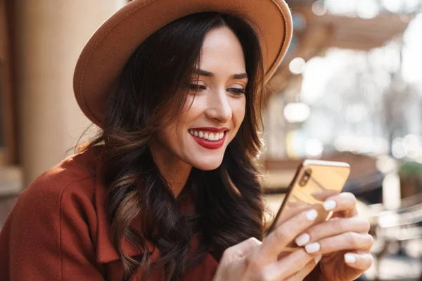 坐在室外街头咖啡馆时 戴着帽子笑着用手机的快乐优雅的成年女性形象 — 图库照片