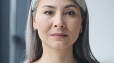 Mutlu gülümseyen olgun bir iş kadınının yakından görüntüsü gözlerini açıyor ve ofisin penceresinin yanındaki kameraya bakıyor.