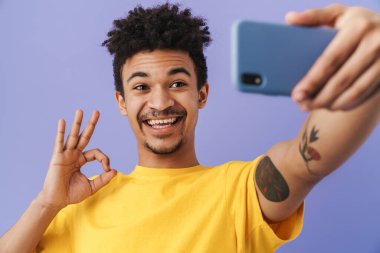 Neşeli Afrikalı Amerikalı bir adamın akıllı telefondan selfie çektiği ve mor arka planda izole edilmiş tamam işareti yaptığı fotoğraf.