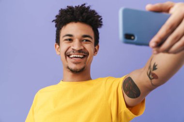 Neşeli Afro-Amerikalı adamın fotoğrafı. Akıllı telefondan selfie çekiyor ve mor arka planda izole bir şekilde gülümsüyor.