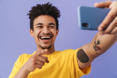 Neşeli Afrikalı Amerikalı bir adamın selfie çektiği ve mor arka planda izole edilmiş akıllı telefonu işaret ettiği fotoğraf.