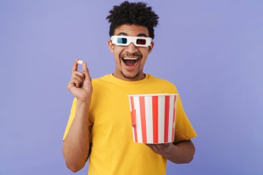 Heyecanlı Afro-Amerikan bir adamın 3 boyutlu gözlüklü fotoğrafı. Film izliyor ve mor arka planda izole edilmiş patlamış mısır yiyor.
