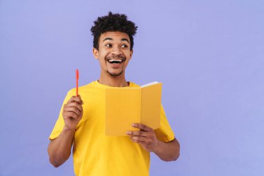 Mor arka planda izole edilmiş kalem ve egzersiz kitabı tutan neşeli Afro-Amerikan adamın fotoğrafı.