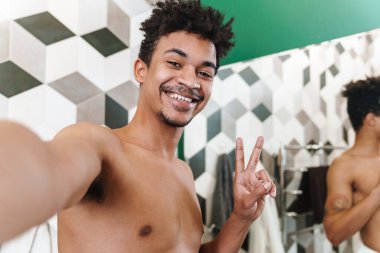 Yarı çıplak gülümseyen Afro-Amerikan adam banyoda selfie çekerken barış işareti yapıyor.