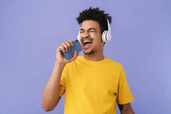 紫色の背景に隔離された携帯電話やワイヤレスヘッドフォンを使用している間に歌う楽しいアフリカ系アメリカ人の男性の写真 — ストック写真