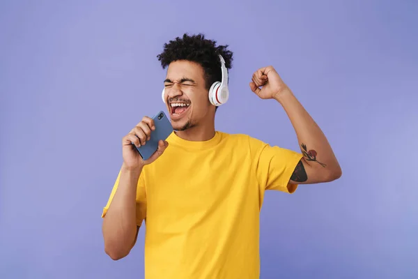 紫色の背景に隔離された携帯電話やワイヤレスヘッドフォンを使用している間に歌う楽しいアフリカ系アメリカ人の男性の写真 — ストック写真