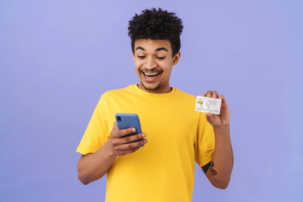 照片中 一个面带微笑的非洲裔美国男人拿着信用卡 用紫色背景隔离的手机 — 图库照片