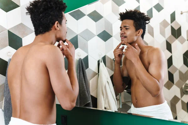 Yarı çıplak, Afro-Amerikan bir adam çenesine dokunuyor ve banyoda aynaya bakıyor.