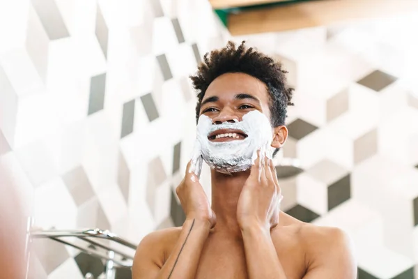 半裸のアフリカ系アメリカ人の男の笑顔の写真顔に剃毛泡で彼の顔に鏡を見てバスルーム — ストック写真