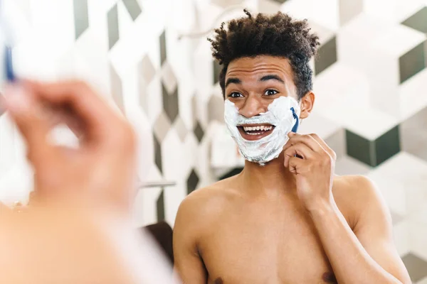 Photo of excited half-naked african american man shaving with foam while looking at mirror in bathroom