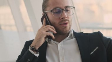 Yakışıklı, genç, sakallı bir iş adamı kapalı bir kafede çalışırken akıllı telefonuyla konuşuyor.